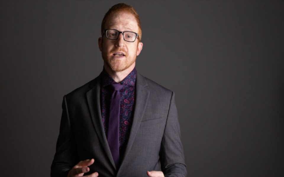 Steve Hofstetter standing in front of a gray back ground while talking