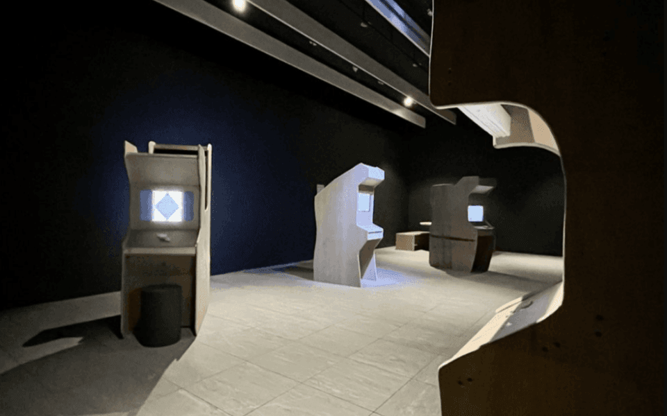 Different retro-style gaming machines lined in a dark hall with white floors. Is an image for the Everest Pipkin Talk at the Anderson Museum of Contemporary Art.